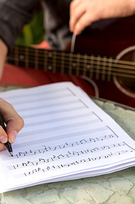 Person skriver noder ned på papir, i baggrunden er en guitar